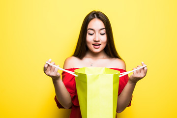 Portrait of a happy pretty asian woman looking inside shopping bag isolated over yellow background