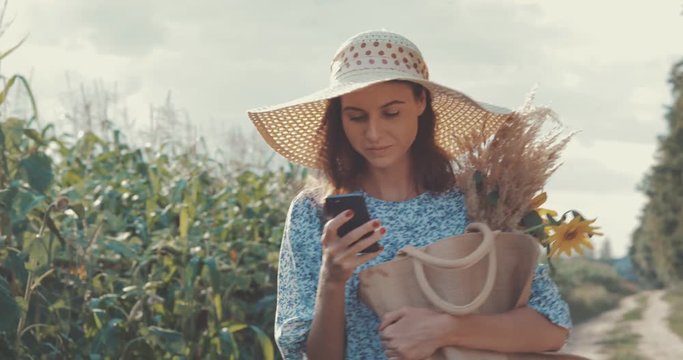 Woman takes selfie on cellphone at sunflower field. Sweet sexy girl in a blue dress and hat walks on a field of sunflowers , lifestyle 4K video shooting by handheld gimbal