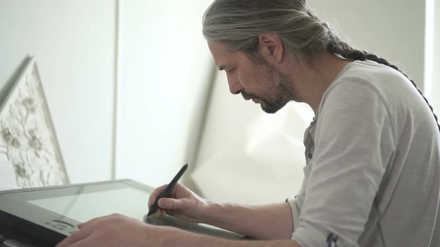 Portrait of senior male digital artist with long grey hair, drawing on the screen of big tablet in his bright studio. Portrait. Profile. Indoors.