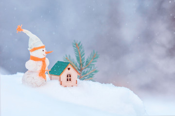 Сhristmas installation with copy space and snowflakes. Funny toy snowman with carrot and orange scarf near the small wooden toy house and green branches of  Christmas tree