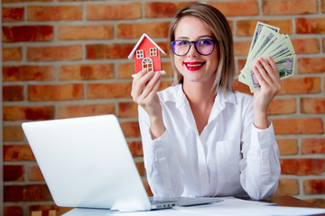 Young realtor with house computer and money in hand on brick wall background