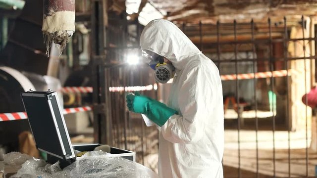 Young ecologist in white protective suit looking at camera. Caucasian guy in hazmat suit and gas mask holding plastic bag in hands. Working day. Indoors.