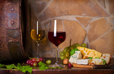 Red and white wine with cheese and grapes beside old cask in wine cellar. Glasses and bottles of wine