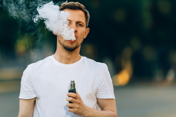 Modern young man with a beard fun Vaporizers Outdoor. Male in white t-shirt vape ecig. dark background, the evening sunset over the city. toned image. - 220881609