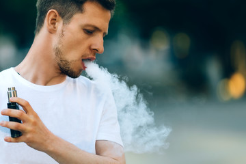 Modern young man with a beard fun Vaporizers Outdoor. Male in white t-shirt vape ecig. dark...