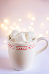 Fototapeta na wymiar White Mug with Hot Chocolate Cocoa Drink and Marshmallows on Top. Glittering Sparkling Garland Bokeh Lights in Background. Soft Pastel Colors. Christmas New Year Festive Romantic Atmosphere.