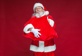 Christmas. A kind smiling Santa Claus is holding a red bag with presents in front of him embracing it. Isolated on red background.