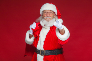 Christmas. Santa Claus in white gloves with a dissatisfied face expression with a bag of gifts behind his back points his index finger into the camera. Isolated on red background.
