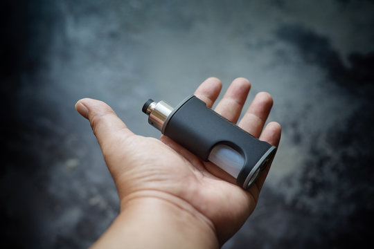 high end unregulated bottom feed squonk box mods with rebuildable dripping atomizer in hand on dark grey texture background, vaping device, vape gear, vaporizer equipment, selective focus