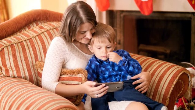 3 years old cute toddler boy sitting with mother in armchair at living room and watching video on screen of mobile phone