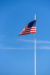 American flag on top white flagpole, flying in the breeze, blue sky background, wispy white cloud, stars and stripes, red, white, and blue
