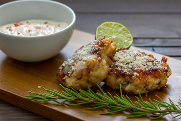 Two shrimp cakes on a cutting board with a lemon garlic dipping sauces lemon wedge and a stem or rosemary on a wooden table.