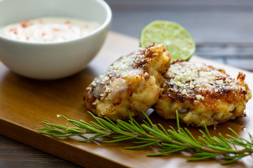 Two shrimp cakes on a cutting board with a lemon garlic dipping sauces lemon wedge and a stem or rosemary on a wooden table.