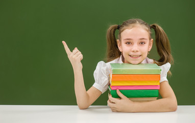 Girl with books points on empty green chalkboard and looking at camera