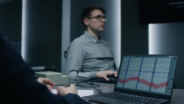 Young Handsome Suspect During Interrogation Undergoes Lie Detector / Polygraph Test, Connected to the Machine He Answers Questions. Computer Shows Measures of Physiological Responds.