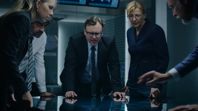 Diverse Team of Government Intelligence Agents Standing Around Digital Touch Screen Table and Tracking Suspect, Senior Officer does Interactive Gesturing. Dark Surveillance Room.