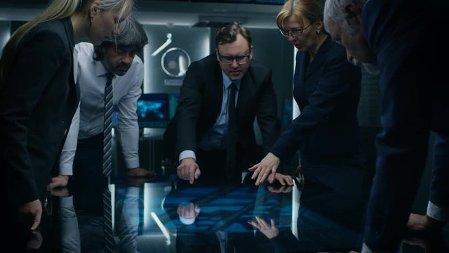 Diverse Team of Government Intelligence Agents Standing Around Digital Touch Screen Table and Tracking Suspect. Big Dark Surveillance Room Full of Computer Screens.