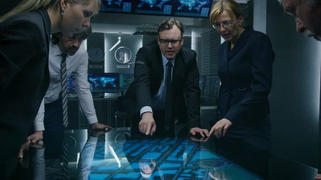 High Angle Shot of Team of Government Intelligence Agents Talking while Standing Around Digital Touch Screen Table and Tracking Suspect. Satellite Surveillance Operation in the Monitoring Room.
