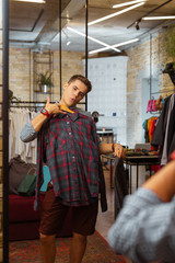 Looking cool. Expressive handsome man holding beautiful shirt and posing with it in front of the mirror while doing shopping