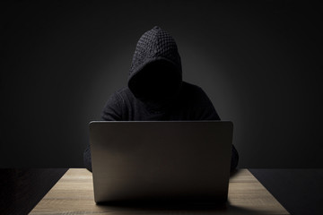 Faceless man in a hooded robe sits at a table with a laptop on a