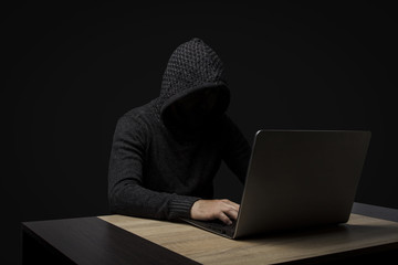 Faceless man in a hooded robe sits at a table with a laptop on a