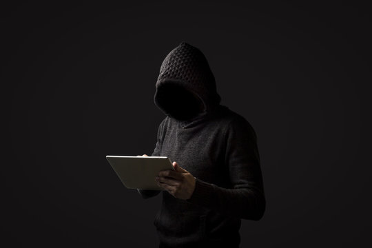 Faceless man in a hoodie with a hood holds a tablet in his hands