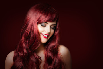 Smiling redhead woman fashion model on dark red background. Makeup, perfect wavy hairstyle