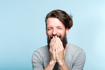 joyful excited happy smiling man covering mouth with hands. portrait of a young bearded guy on blue...