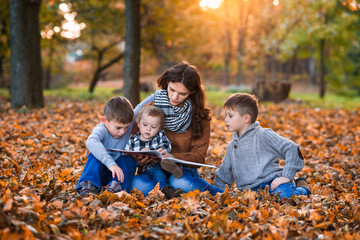 A young beautiful mother reads a book with fairy tales for her three sons in the autumn park. Mom with children in the autumn background.