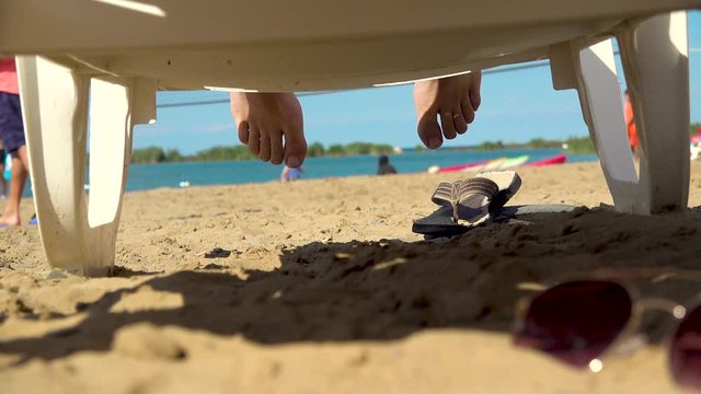 View from under a lounger as sun worshipper relaxes on a beach, feet lazily playing, with people playing anonymously in background