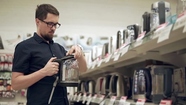 Householder is examining sample of electric tea kettle in a trading area in supermarket. He is looking on it, opening and closing cover