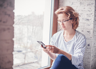 Young attractive woman  in glasses sits on a windowsill with a mobile phone smartphone in hands, reading and education using applications for the phone