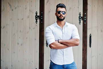 Stylish tall arabian man model in white shirt, jeans and sunglasses posed at street of city. Beard...