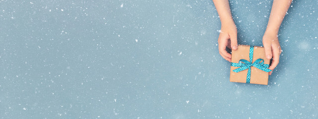 Child hands holding one gift box wrapped in kraft paper tied with blue ribbon in polka dots on blue grey background. Top view, place for text. Holiday concept, banner for site.