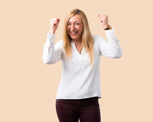 Middle age blonde woman with white shirt celebrating a victory in winner position on isolated ocher...