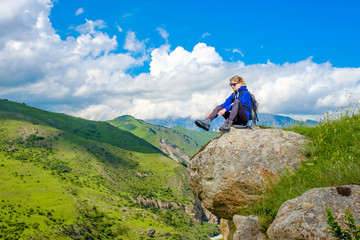 Fototapeta premium A girl with a backpack is sitting on a rock. Mountain landscape. Mountain climbing in the Caucasus.