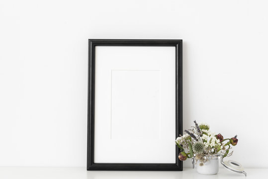 Black a4 portrait frame mockup with small bouquet of flowers in small white pot on white wall background. Empty frame, poster mock up for presentation design.