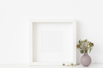 White square portrait frame mockup with a small bouquet of dried flowers in a lilac small vase on white wall background. Empty frame, poster mock up for presentation design. 