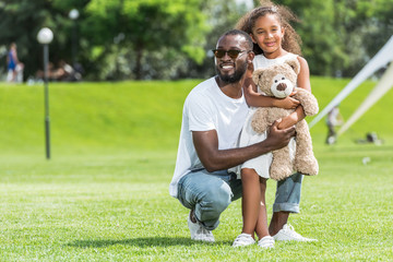 smiling african american father squatting and hugging daughter with teddy bear in park
