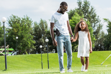 Fototapeta premium african american cop with gun in holster and daughter holding hands and walking at amusement park