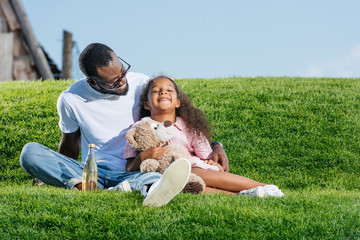 african american father and smiling daughter sitting on hill with soda and teddy bear at amusement park