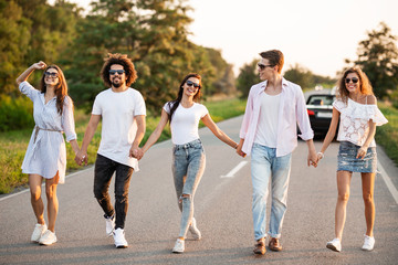 Obraz premium Young happy guys and girls are holding hands and walking on the road on a sunny day