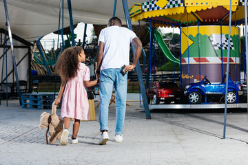 rear view of african american policeman walking with daughter and holding police badge at amusement park