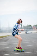 A beautiful blond girl wearing checkered shirt, cap and denim shorts is longboarding while stretching out her hands. Sport and cool style.