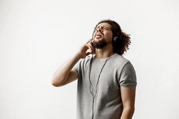 A curly-headed handsome man wearing a gray T-shirt and ripped jeans is standing and listening to music in the headphones and looking upwards with his finger on the ear over the white background.