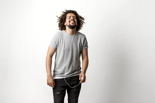 A curly-headed handsome man wearing a gray T-shirt and ripped jeans is listening to music in the earphones with a blissful expression on the face over the white background.