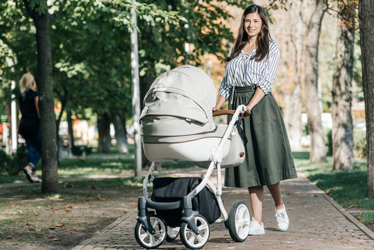 beautiful mother walking with baby stroller in park and looking at camera