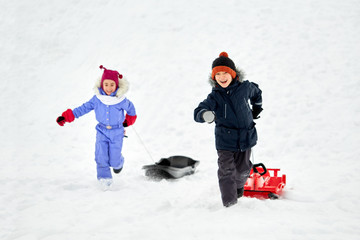 childhood, leisure and season concept - happy little kids in winter clothes with sled having fun outdoors