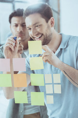 smiling young businessman writing on sticky notes while focused colleague standing behind