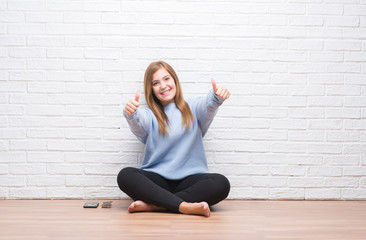 Young adult woman sitting on the floor in autumn over white brick wall approving doing positive gesture with hand, thumbs up smiling and happy for success. Looking at the camera, winner gesture.